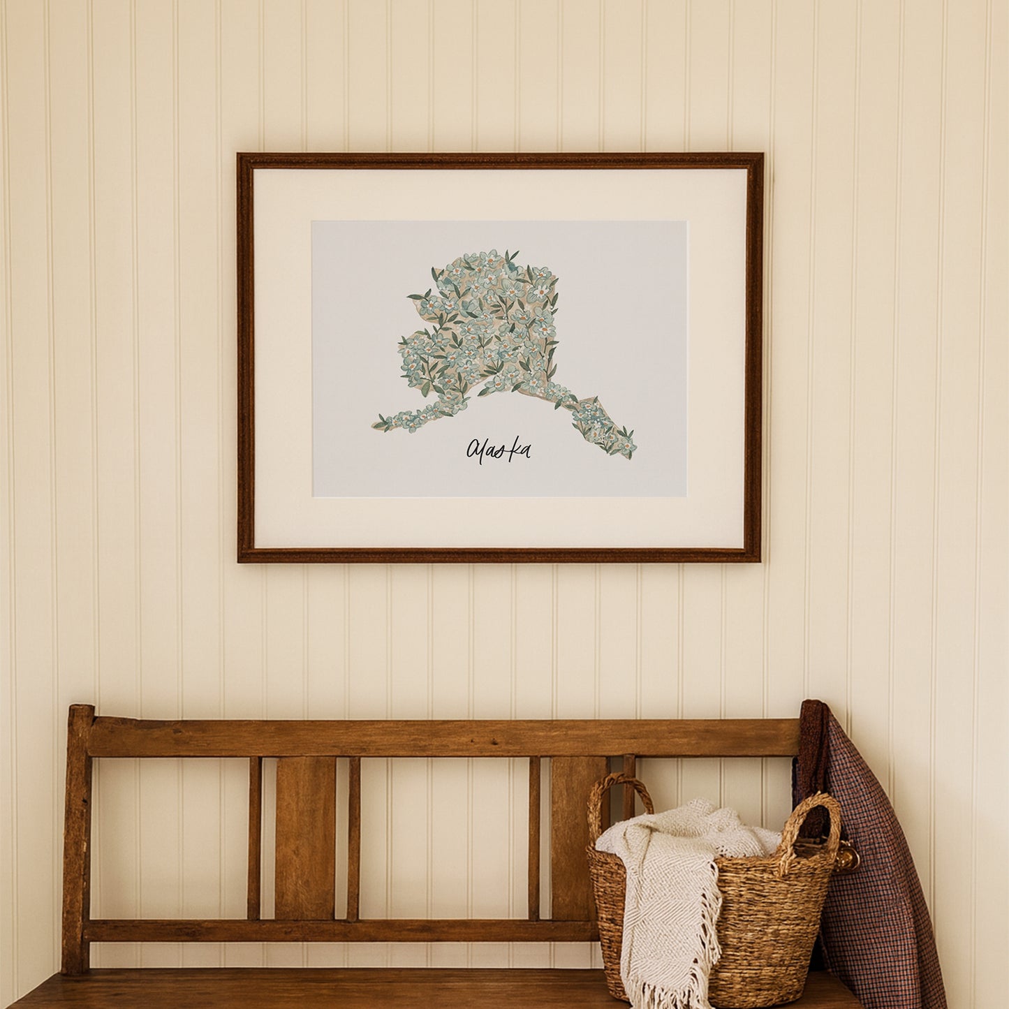 Framed Alaska state flower watercolor artwork on a wall above a wooden bench with a basket and blanket.