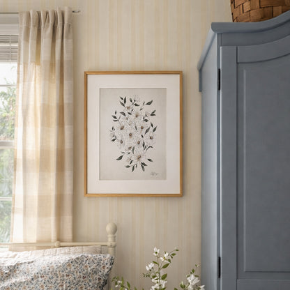 Framed April daisy birth flower watercolor art print in soft neutral tones on a wall next to a window with curtains and a blue gray cabinet.