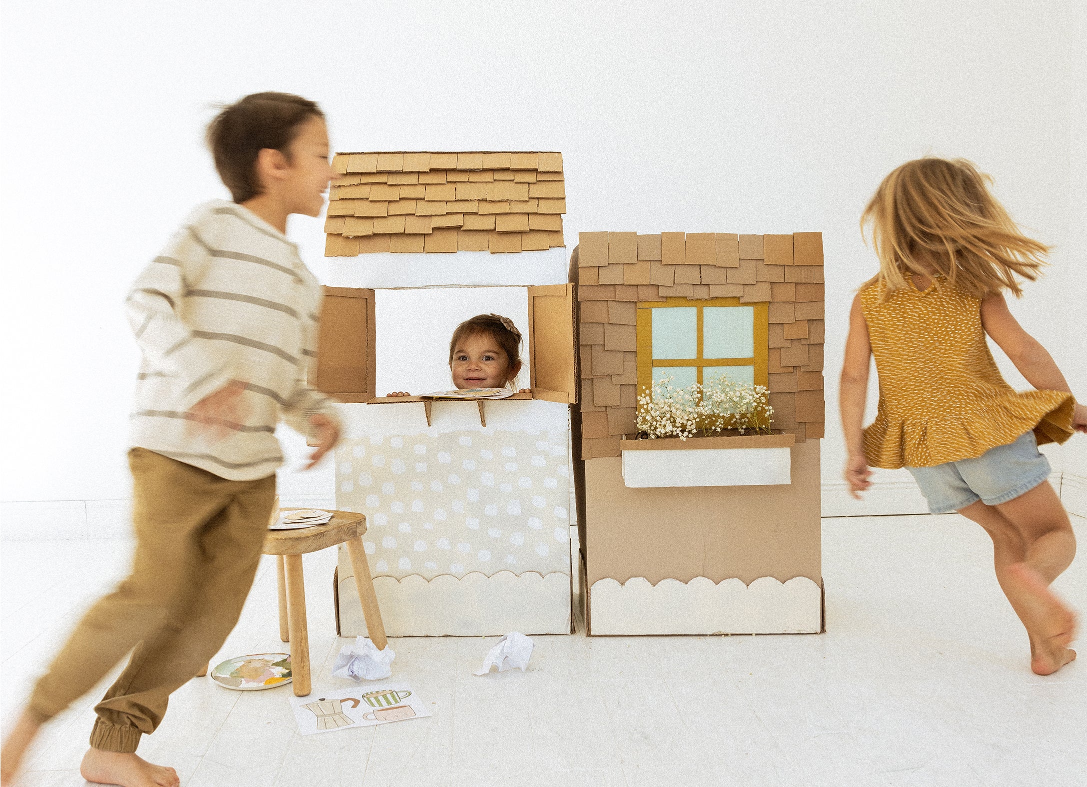 Children playing with a cardboard house on a white floor.