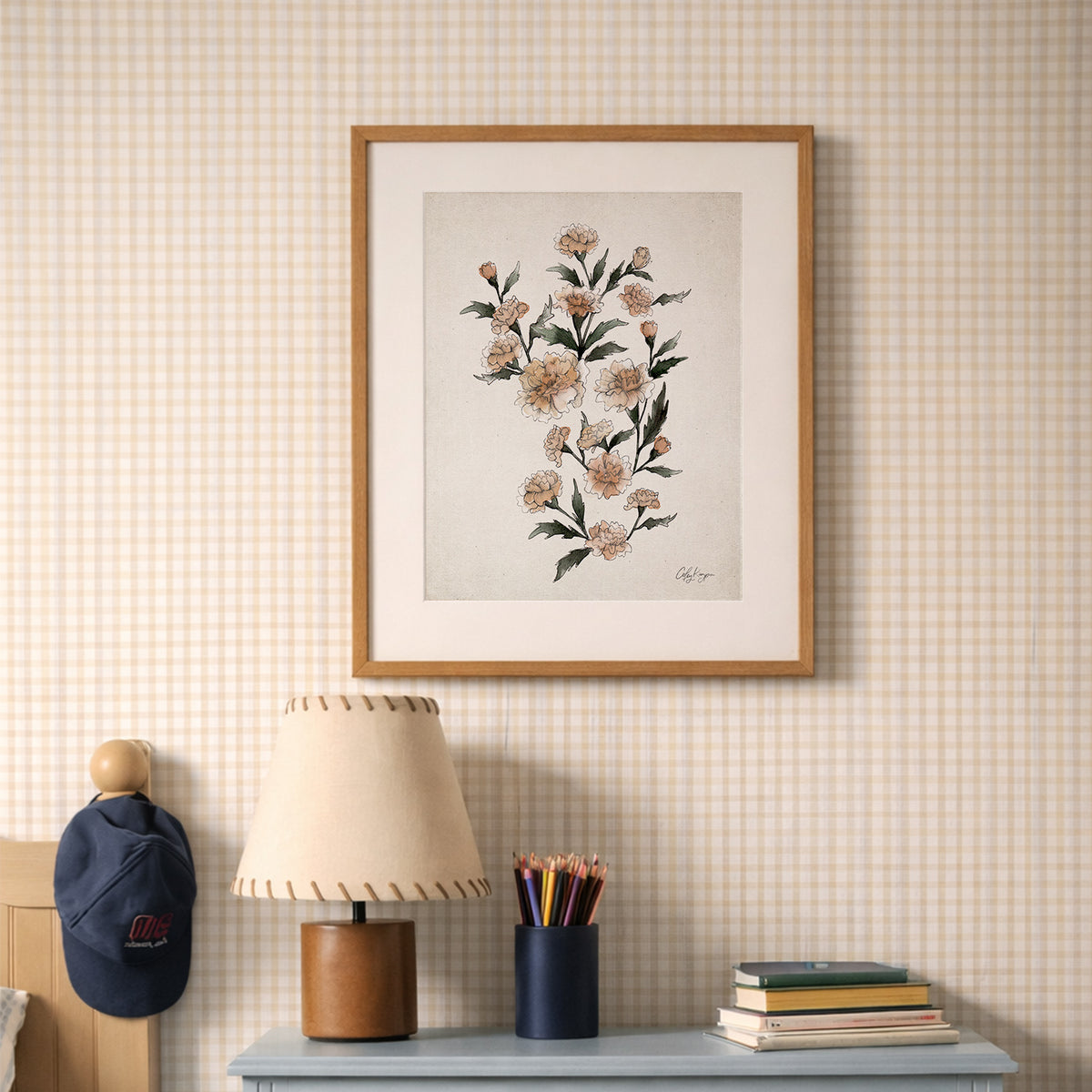 Marigold birth flower wall art – October watercolor art print by Coley Kuyper artwork on a wall with a lamp, books, and stationery items on a shelf below.